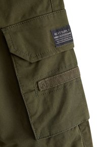 Tasca cargo in tessuto verde con patta e etichetta cucita con la scritta "Next Supply Authentic Craft Durable Goods."