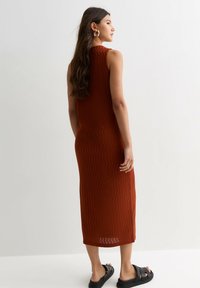 Robe sans manches de couleur rouille avec une texture ondulée, tombant jusqu'au mollet. Portée avec des sandales noires, mettant en avant un design minimaliste.