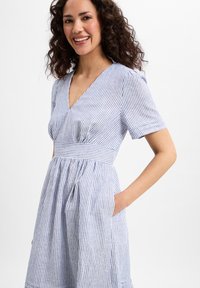 Gestreiftes Kleid mit V-Ausschnitt, kurzen Ärmeln, tailliertem Oberteil und schwingendem Rock mit seitlichen Taschen. Blau-weiße Farben. Weiche Oberfläche.