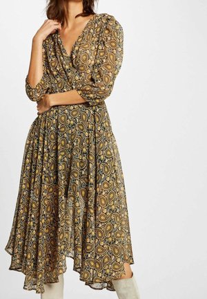 Kobieta ubrana w midi sukienkę z długim rękawem w orientalny wzór paisley w odcieniach brązu i złota, z białymi kozakami, pozująca z jedną ręką przy twarzy.