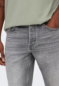 Jeans de denim gris con un ajuste ajustado, que presentan un sutil desgaste y un diseño de cinco bolsillos. Combinados con una camiseta de algodón verde claro.