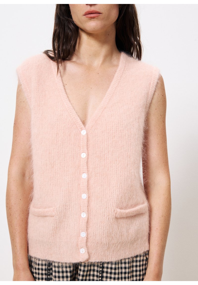 Gilet sans manches rose en matériau doux et texturé, avec un col en V, sept boutons blancs et deux poches avant. Pantalons à carreaux noir et blanc visibles.