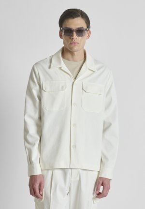 Uomo con giacca color crema con due tasche sul petto, pantaloni coordinati, camicia beige e occhiali da sole scuri su sfondo semplice.