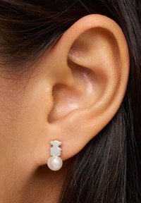Zbliżenie ucha z srebrnym kolczykiem w kształcie misia, zakończonym okrągłą białą perłą.