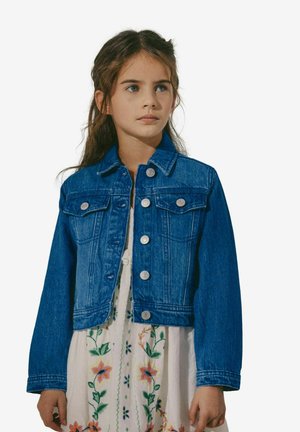 Ung pige med langt hår bærer blå denimjakke over hvid kjole med farverig blomsterbroderi, stående mod ensfarvet baggrund.