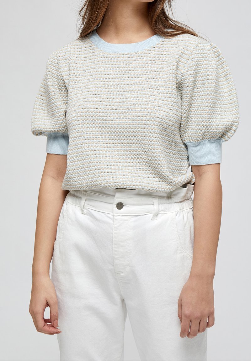 Femme portant un pull bleu clair texturé à manches courtes bouffantes et un pantalon blanc taille haute.