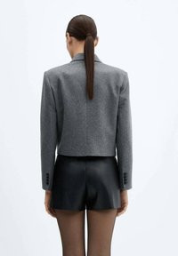 Veste grise courte en laine, avec un col cranté, des manches longues ornées de boutons, associée à un short en cuir noir.