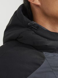 Giacca nera con cappuccio in un tessuto texture con accenti opachi e lucidi, che mostra pannelli a contrasto e coulisse regolabili.