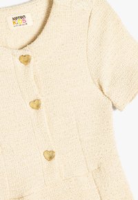 Crème-kleurige kinderkleed gemaakt van gestructureerde stof, met korte mouwen en hartvormige gouden knopen.