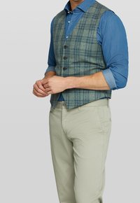 Grön rutmönstrad väst över en blå skjorta med knappar, kompletterad med ljusbeiga byxor. Har fem knappar och en skräddarsydd passform.