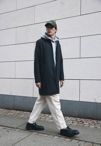 Dunkelgrauer Wollmantel, grauer Hoodie, crèmefarbene Cordhosen, schwarze Schuhe mit dicken Sohlen. Model geht vor einem hellen Steinfond.