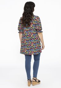 Tunique florale en tissu noir avec des motifs de papillons colorés, manches bouffantes et ourlet arrondi, assortie à un jean skinny bleu et des sandales compensées.