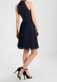 Námořnicky modré šaty na krk s texturovaným živůtkem a skládáním na sukni. Doplněné černými sandály na vysokém podpatku s detaily na kotníku.