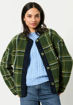 Ung kvinde iført en grøn ternet jakke med knapper over en lyseblå kabelstrikket sweater og blå jeans, står med hænderne i lommerne.