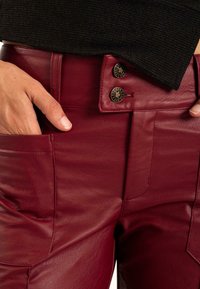 Los pantalones de cuero rojo presentan una textura suave, un cierre de dos botones y bolsillos laterales. El diseño incluye costuras ajustadas para un look entallado.