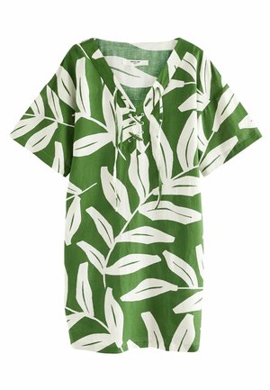 Personne portant une robe verte à manches courtes avec un motif de feuilles blanches, debout dans une eau peu profonde en éclaboussant avec un pied.