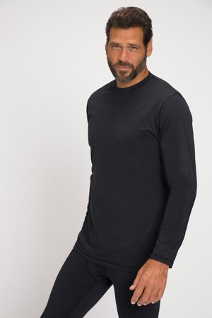 Mann mit Bart, der ein eng anliegendes schwarzes Langarmshirt und schwarze Hose trägt, steht vor einem schlichten hellen Hintergrund.