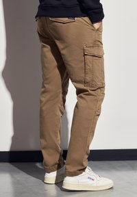 Béžové cargo kalhoty se bočními kapsami, zúženými nohavicemi a měkkou texturou látky, spárované s bílými teniskami s zelenými akcenty.