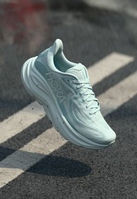 Gaiši zila skriešanas apava ar elpojošu acs virsu, polsterētu apkakli, bieza balta zole un HOKA logotipu uz sāna. Peldoša virs teksturētas virsmas.