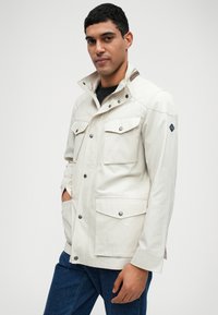 Chaqueta de color beige claro, con botones, cuatro bolsillos frontales, cuello alto y cierres a presión. Textura suave con un diseño moderno.