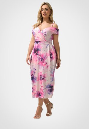 Kvinde iført en pink og lilla blomsterprintet off-shoulder midi-kjole med tynde stropper og gyldne højhælede sandaler på en hvid baggrund.