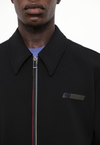 Veste zippée noire avec un col pointu, tissu texturé et un logo en patch. Présente un détail de fermeture éclair multicolore sur le devant.