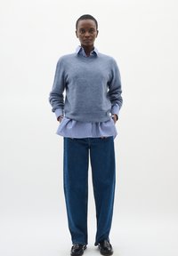 Svetlo modra volnena pulover z V-izrezom, prekrite s modro in belo prugasto srajco z nabranim robom, v kombinaciji s temno modrimi širokimi hlačami.