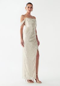 Večerna obleka iz smetanasto belega sequina z ramen brez ramen, prilagojenim krojem, stranskim razporkom in elegantnimi nabranim tkaninami. Zlati petki dopolnjujejo videz.