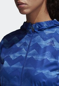 Veste zippée bleue arborant un motif en chevrons, fabriquée à partir d'un tissu lisse et léger avec une finition texturée. Conception à capuche avec bord élastique.