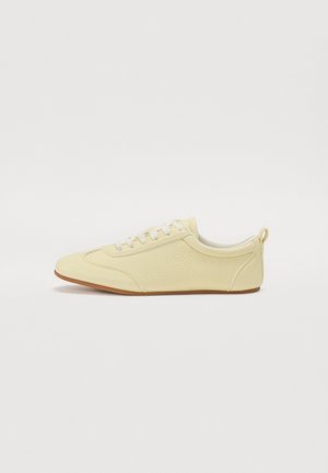 Licht beige lage sneaker met witte veters, gestructureerd materiaal en een zool van gum, zijaanzicht op een witte achtergrond.