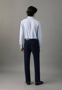 Svetlomodrá košeľa s dlhým rukávom a manžetami na gombíky, v kombinácii s tmavomodrými nohavicami. Tkanina košele vyzerá hladko; nohavice majú prispôsobený strih.