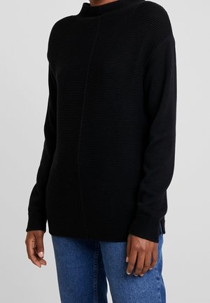 Personne portant un pull en maille noire texturée à manches longues et un jean bleu, debout devant un fond gris clair uni.
