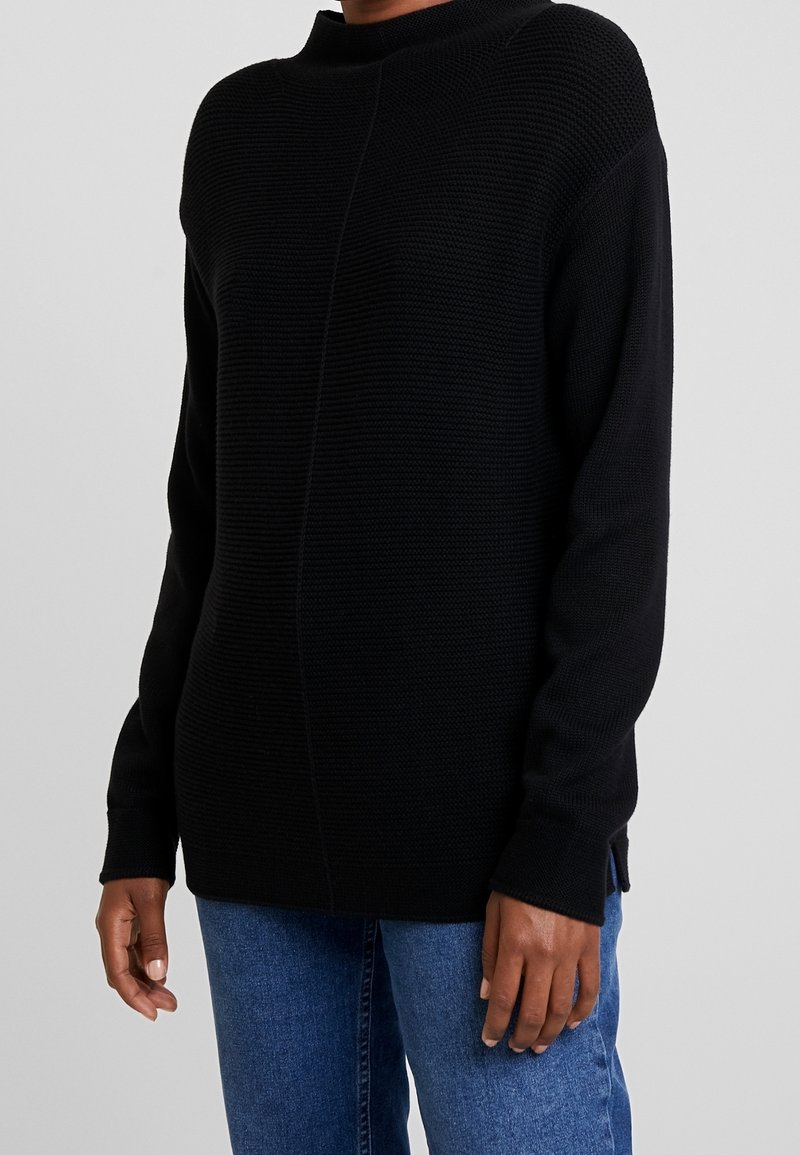 Personne portant un pull en maille noire texturée à manches longues et un jean bleu, debout devant un fond gris clair uni.