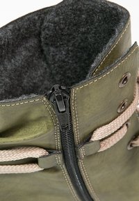 Gros plan sur une botte en cuir verte avec une fermeture éclair noire, des lacets beige, des œillets en métal et une doublure intérieure grise et duveteuse.