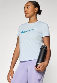 Lyseblå treningstrøye med et turkist Nike swoosh-logo, kombinert med lavendel tights og en svart vannflaske. Glatt tekstur.