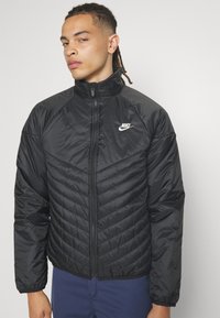 Giacca invernale nera con colletto alto, caratterizzata da un motivo trapuntato sul corpo e una chiusura con zip. Logo Nike sul lato sinistro del petto.