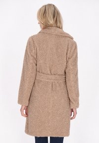 Cappotto fuzzy beige con cintura, maniche lunghe e collo. Texture morbida e lunghezza media, progettato per calore e comfort.