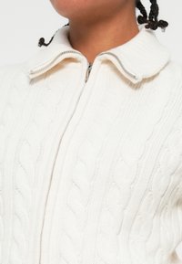 Cardigan en tricot couleur crème avec un motif en relief, doté d'une fermeture éclair et d'un col arrondi. Tissu doux et texturé avec des accents côtelés.