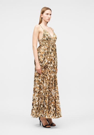 Γυναίκα που φοράει μακρύ φόρεμα σε λεοπάρ σχέδιο με πιέτες, λεπτές τιράντες και μαύρα πέδιλα με ψηλό τακούνι, σε λευκό φόντο.