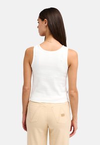 Débardeur blanc sans manches avec un col arrondi, fabriqué à partir d'un tissu extensible, associé à un pantalon beige clair avec des poches en forme de triangle.