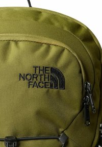 Mochila verde oliva hecha de un tejido duradero que presenta un destacado logo negro, superficie texturada y compartimentos con cremallera con herrajes negros.