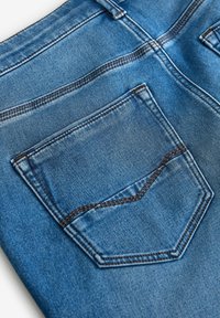 Primer plano de unos pantalones vaqueros azul denim que muestran el bolsillo trasero con costura marrón detallada y el pasador del cinturón sobre un fondo blanco.