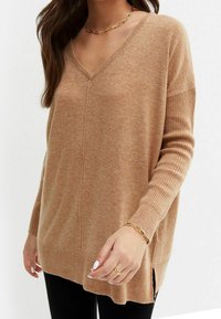 Vrouw in een camelgekleurde V-hals trui met geribbelde mouwen, zwarte broek, gouden kettinghalsketting, armband en ring, met witte nagellak.