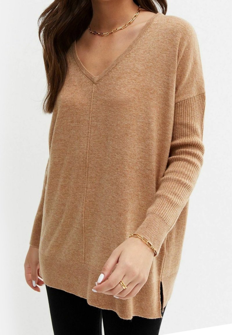 Vrouw in een camelgekleurde V-hals trui met geribbelde mouwen, zwarte broek, gouden kettinghalsketting, armband en ring, met witte nagellak.