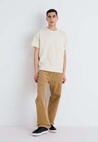 T-shirt crème à manches courtes associé à un pantalon en velours côtelé beige. Des sneakers noires avec des semelles blanches complètent la tenue. Textures lisses évidentes.