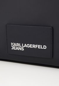 Mustavesi muovi nahasta valmistettu laukku, jossa on suorakulmainen etiketti, jossa on kohokirjoituksella valkoinen teksti "KARL LAGERFELD JEANS." Niukasti muotoiltu ja sileä pinta.