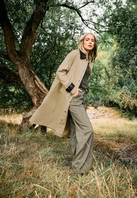 Cappotto impermeabile beige con colletto nero, abbinato a pantaloni larghi a quadri verdi, ambientato in un contesto naturale all'aperto.