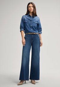 Camicia di denim blu medio con due tasche sul petto, abbinata a jeans a gamba larga blu. Una cintura beige mette in evidenza la vita. Tacchi alti neutri.