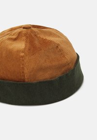 Cordfløyel lue med oransje overdel og mørkegrønn ribbet mansjett. Rund form, myk tekstur med vertikale ribber.