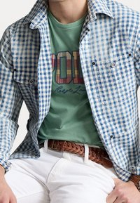 Blue and white gingham shirt with a buttoned front over a green t-shirt featuring a multicolored logo, paired with white pants and a brown braided belt.